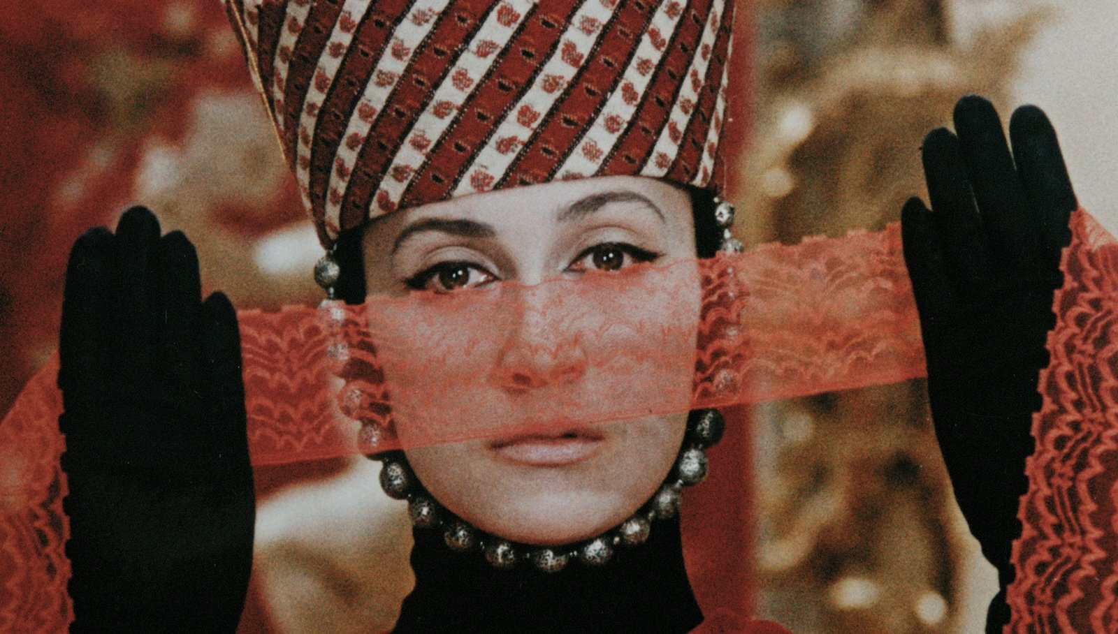 A person in an elaborate red and white hat that looks more like a royal crown holds a gossamer red scarf up to her face, wearing black gloves