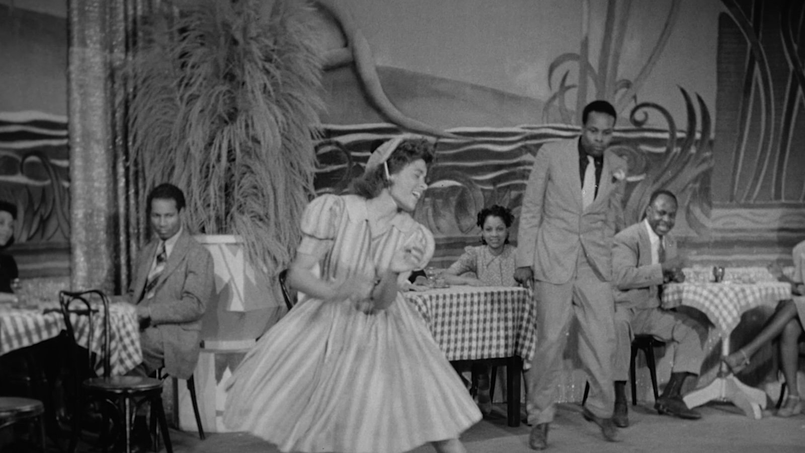 Black and white image from the 1940s of a woman in a dress dancing in a cafe while men look at her; one in a suit is standing, the others are sitting.