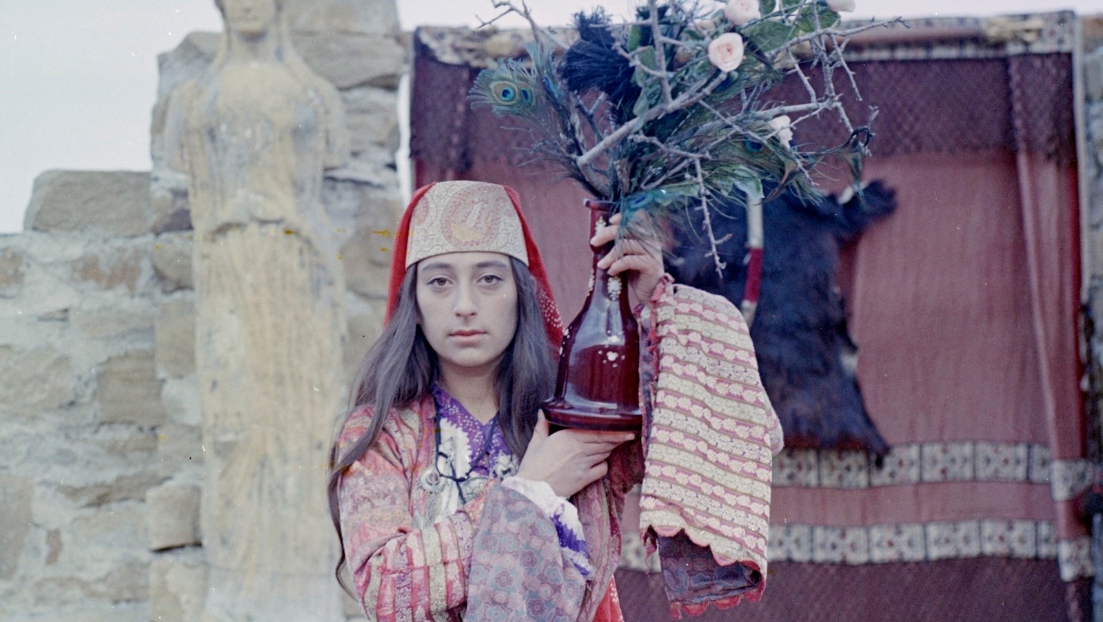 A person with long dark hair and dressed in colorful patterns and a middle-eastern style hat stands in front of a curtain draped outdoors on a door opening, looks ahead with a serious expression while holding up a red glass bottle full of flowers