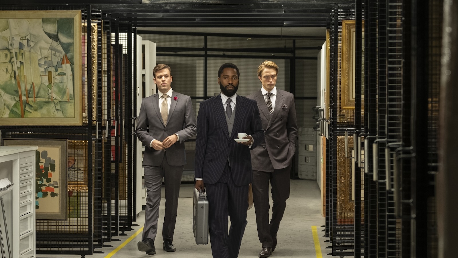 Three men in suits walk towards a camera in a storage room filled with paintings. The man in front carries a teacup and a suitcase.