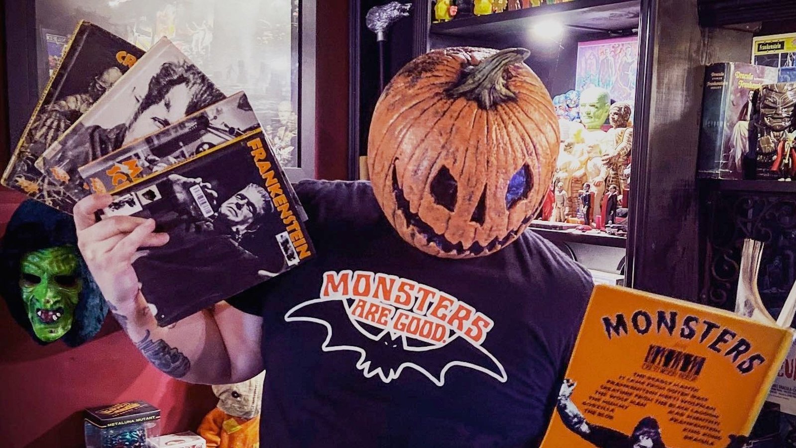 A figure wearing a Jack O Lantern mask stands in front of a shelf of monster memorabilia and holds monster movie magazines, one featuring Frankenstein's monster and another featuring King Kong.