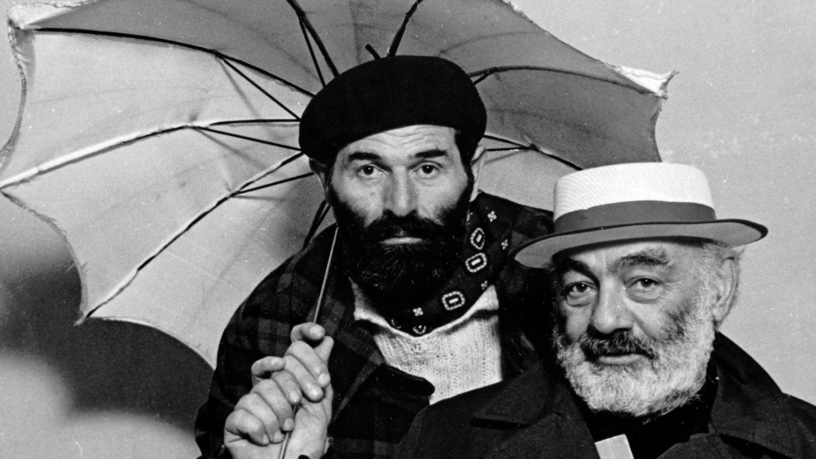 Two bearded, hatted men, one with a gray beard the other with a brown beard stand together looking at us; the younger one in the back holds an open umbrella.