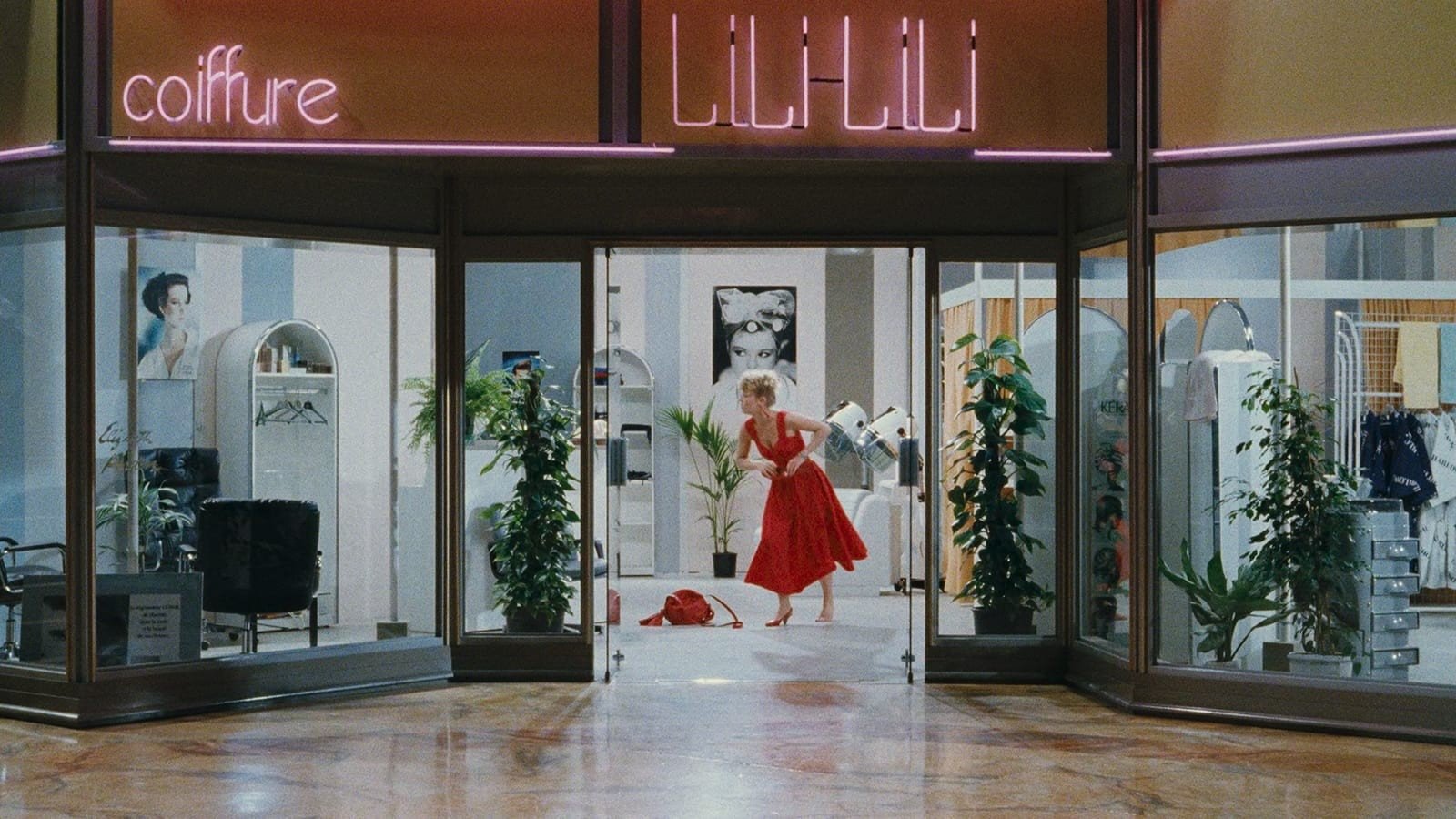 A woman in a red dress and heels stands framed by a hair salon's doorway in an indoor shopping mall with the word "coiffure" in neon above the door to the left
