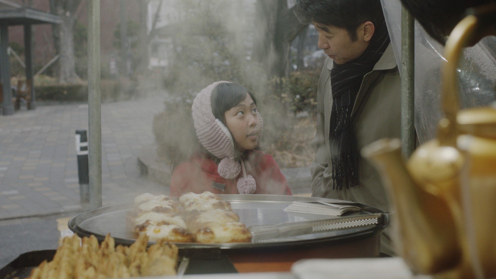 A little girl in a pink winter hat with pom poms looks up at an older man, maybe her father, while standing in front of a food cart with a tray of pastries on it