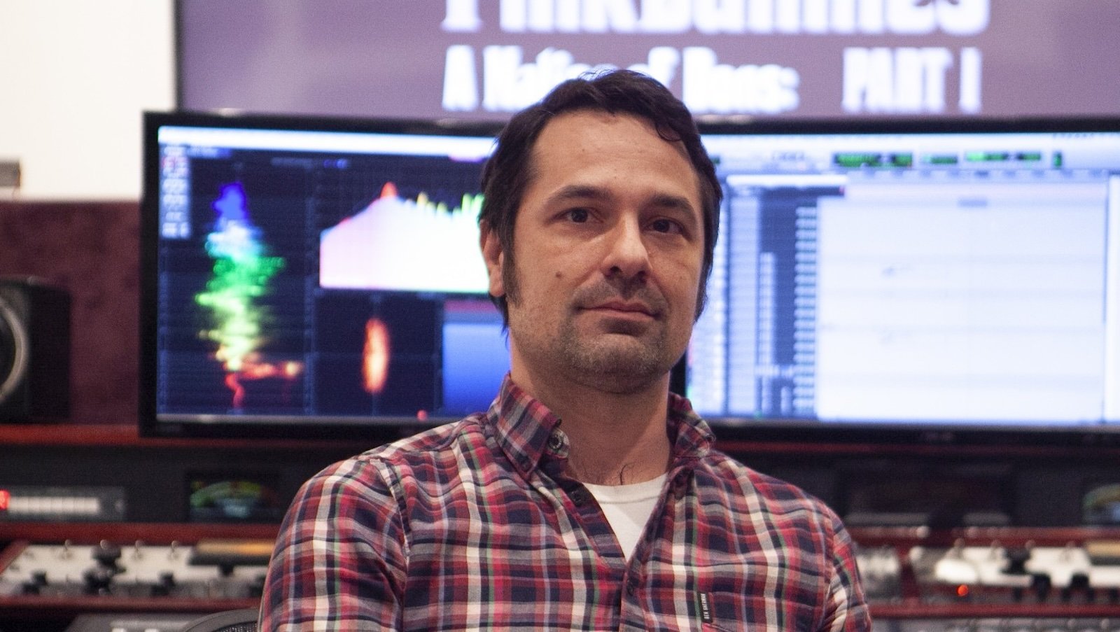 A dark-haired man wearing flannel shirt and a serious expression looks directly at us while sitting in front of two large computer monitors displaying film editing software